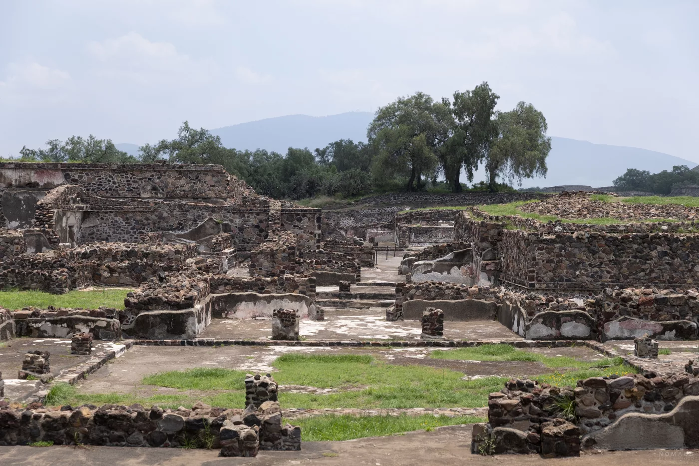 Ruinen einer antiken Wohnanlage in Teotihuacán mit Baumgruppen und grünen Hügeln im Hintergrund.