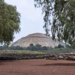 Die Sonnenpyramide von Teotihuacán, eingerahmt von zwei Bäumen im Vordergrund, mit Blick über rote Erde und grüne Vegetation.
