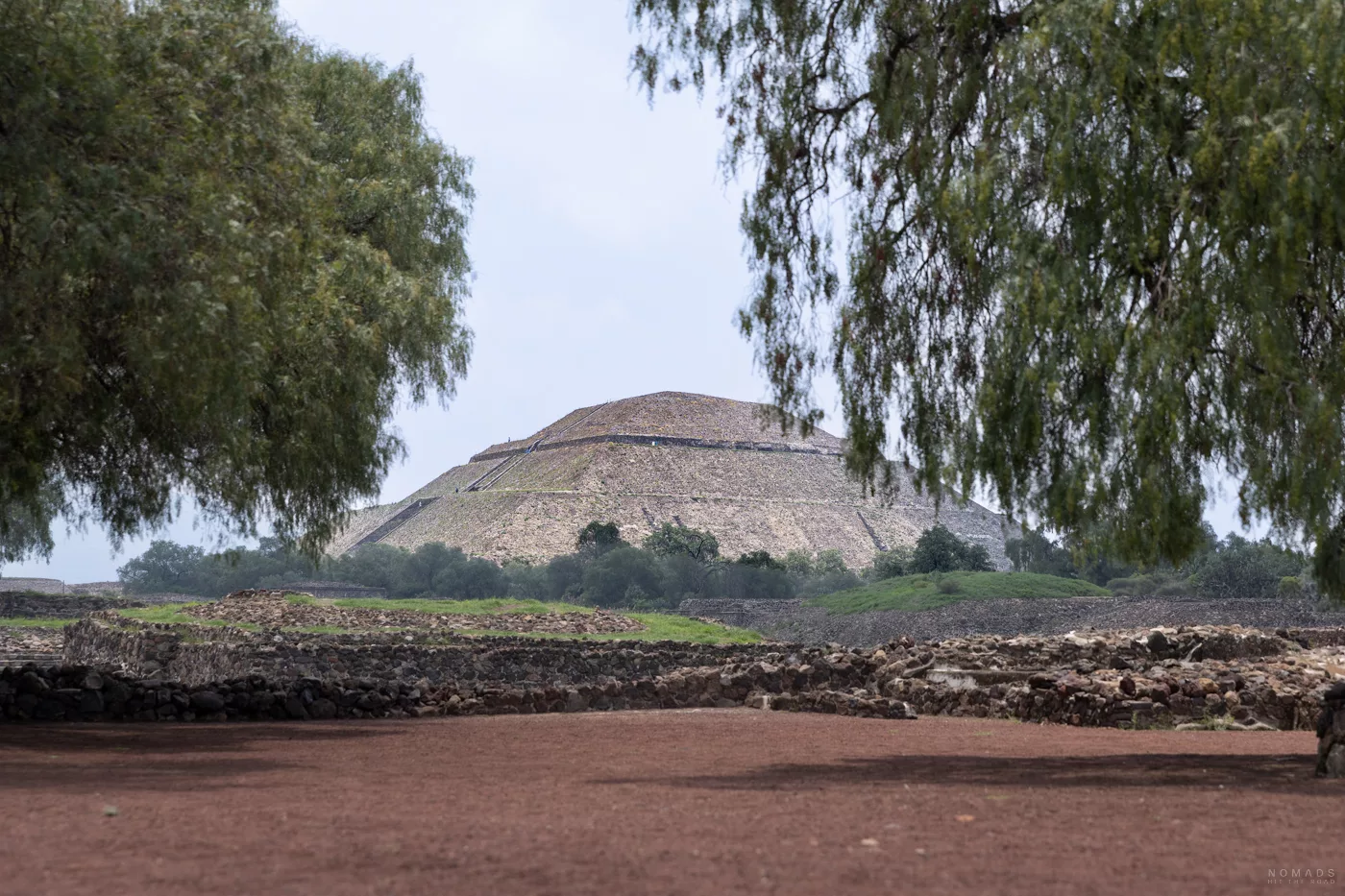 Die Sonnenpyramide von Teotihuacán, eingerahmt von zwei Bäumen im Vordergrund, mit Blick über rote Erde und grüne Vegetation.