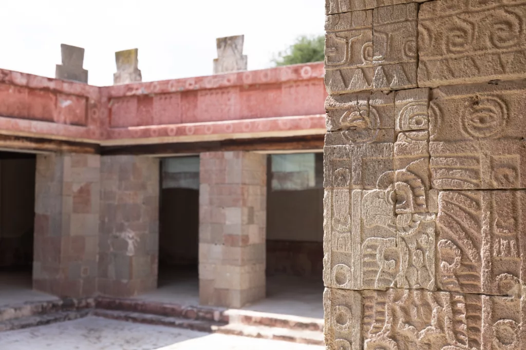 Detailaufnahme eines kunstvoll verzierten Steinsäulenfragments mit Spiralmotiven und stilisiertem Vogelrelief im Palast des Quetzalpapálotl in Teotihuacán; im Hintergrund sind mehrere Räume des rechteckigen Innenhofs mit bemalten roten Friesen sichtbar.
