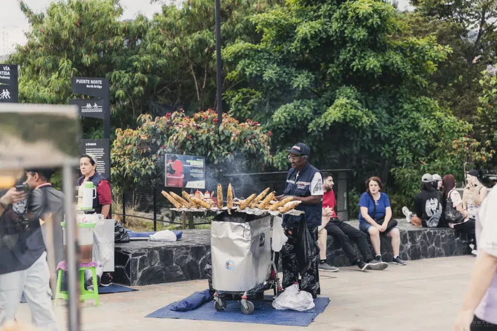 Straßenverkäufer in Medellín grillt Maiskolben an einem mobilen Stand, während Menschen im Hintergrund sitzen und sich unterhalten.