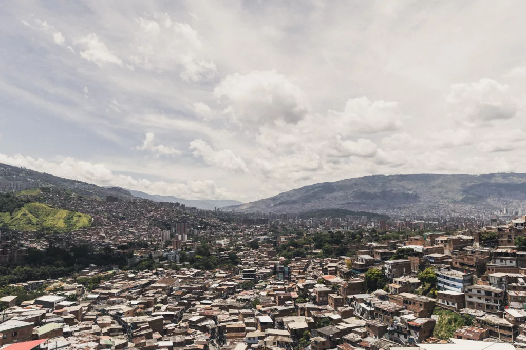 Panorama von Medellín mit dicht bebauten Häusern im Vordergrund, modernen Hochhäusern im Tal und den umliegenden grünen Bergen unter bewölktem Himmel.