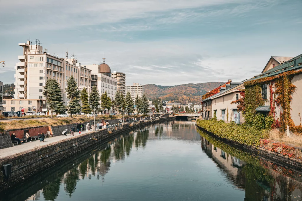 Der malerische Otaru-Kanal mit bewachsenen Lagerhäusern und Promenade, umgeben von herbstlicher Kulisse und Bergen im Hintergrund.
