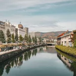 Der malerische Otaru-Kanal mit bewachsenen Lagerhäusern und Promenade, umgeben von herbstlicher Kulisse und Bergen im Hintergrund.