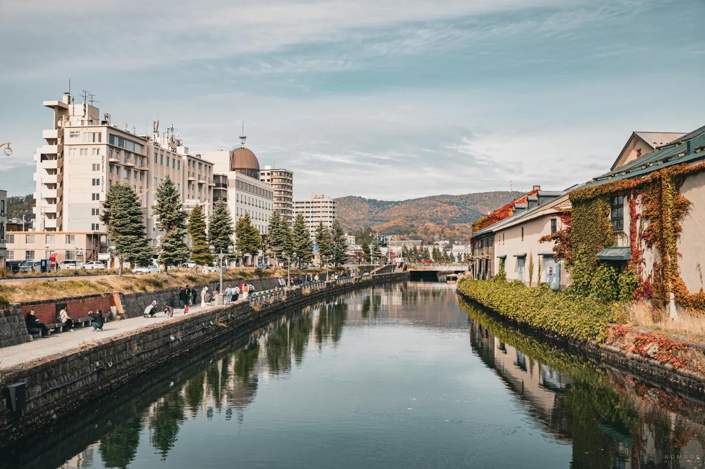 Der malerische Otaru-Kanal mit bewachsenen Lagerhäusern und Promenade, umgeben von herbstlicher Kulisse und Bergen im Hintergrund.