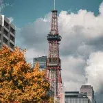 Sapporo TV Tower an einem herbstlichen Tag, umgeben von buntem Laub und moderner Stadtarchitektur unter dramatischem Himmel.