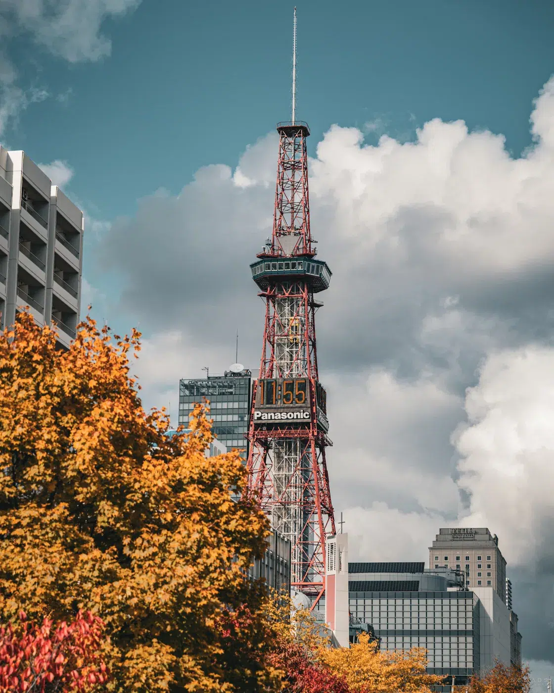 Sapporo TV Tower an einem herbstlichen Tag, umgeben von buntem Laub und moderner Stadtarchitektur unter dramatischem Himmel.