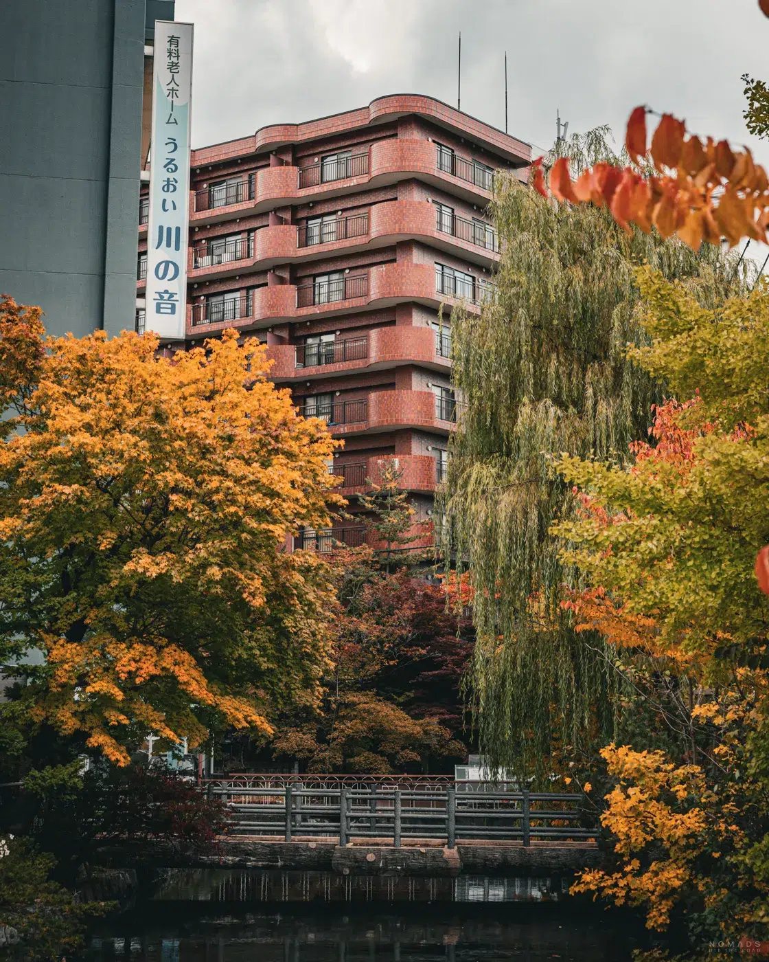 Ruhige Szene im Herzen von Sapporo mit herbstlich gefärbten Bäumen, einer kleinen Holzbrücke und einem roten Backsteingebäude im Hintergrund.