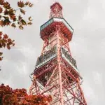 Detailaufnahme des roten Sapporo Fernsehturms mit Aussichtsplattform, umrahmt von herbstlich gefärbten Ahornblättern.