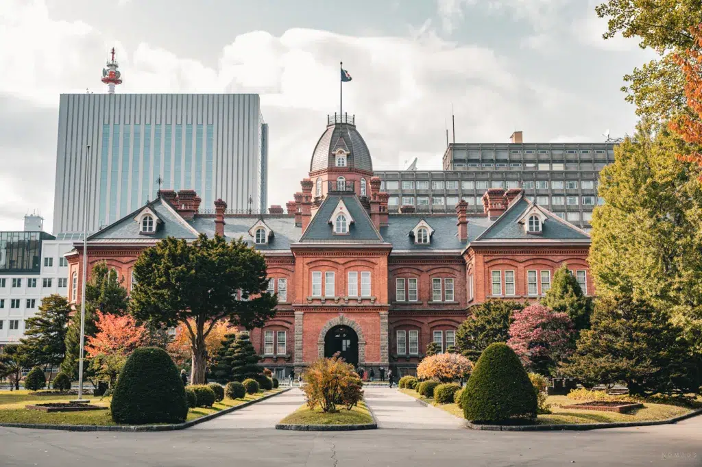 Backsteingebäude der ehemaligen Regierung von Hokkaido in Sapporo, umgeben von herbstlich gefärbtem Garten und moderner Stadtarchitektur im Hintergrund.