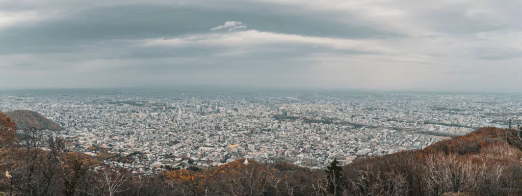 Weitwinkelblick über die Stadt Sapporo vom Aussichtspunkt des Mount Moiwa – herbstliche Hügel im Vordergrund, dichtes Stadtbild bis zum Horizont.