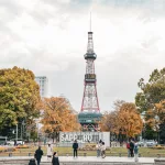 Sapporo Fernsehturm im Herbst, gesehen vom Odori Park aus, umgeben von bunten Bäumen und Spaziergängern vor dem SAPPORO-Schriftzug.