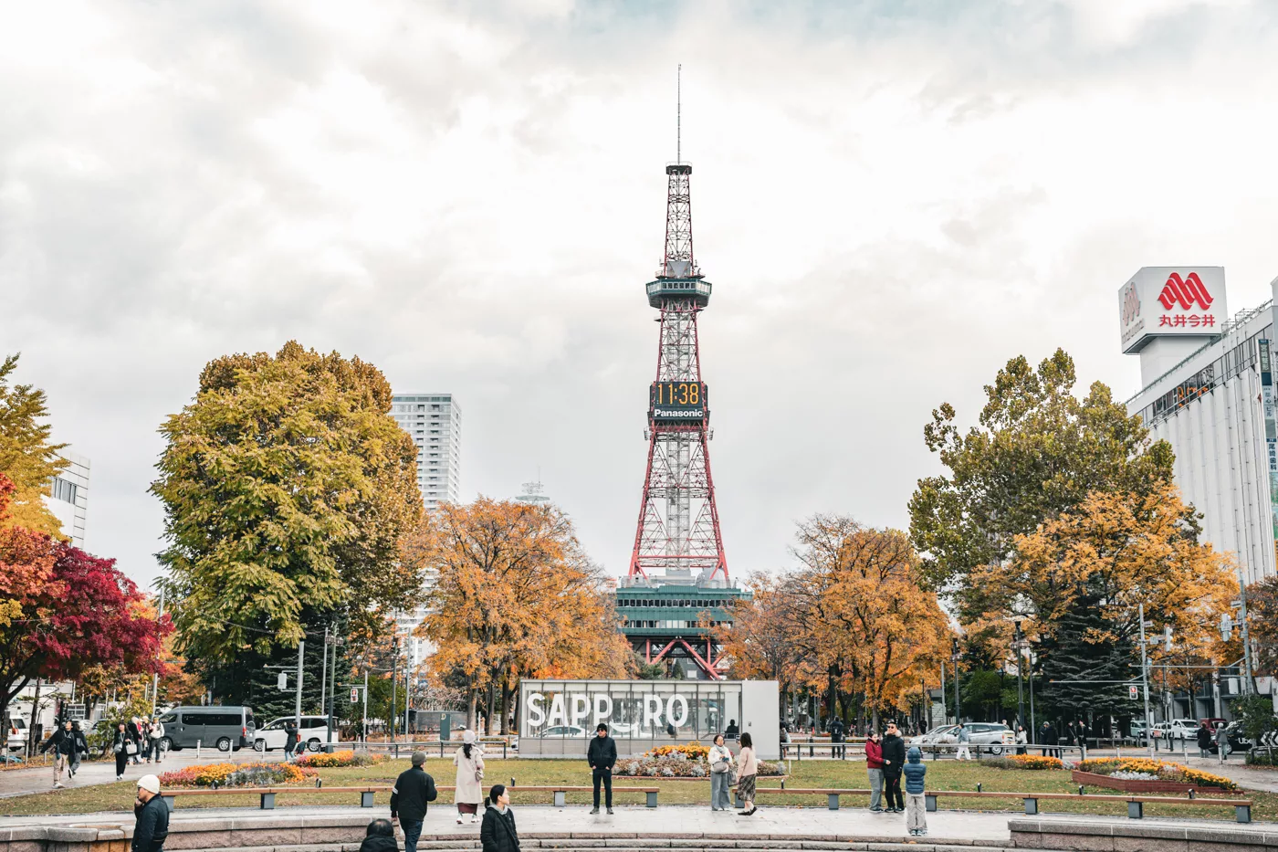 Sapporo Fernsehturm im Herbst, gesehen vom Odori Park aus, umgeben von bunten Bäumen und Spaziergängern vor dem SAPPORO-Schriftzug.