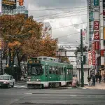 Grüne Straßenbahn in der Innenstadt von Sapporo im Herbst, umgeben von buntem Laub und japanischen Reklametafeln.