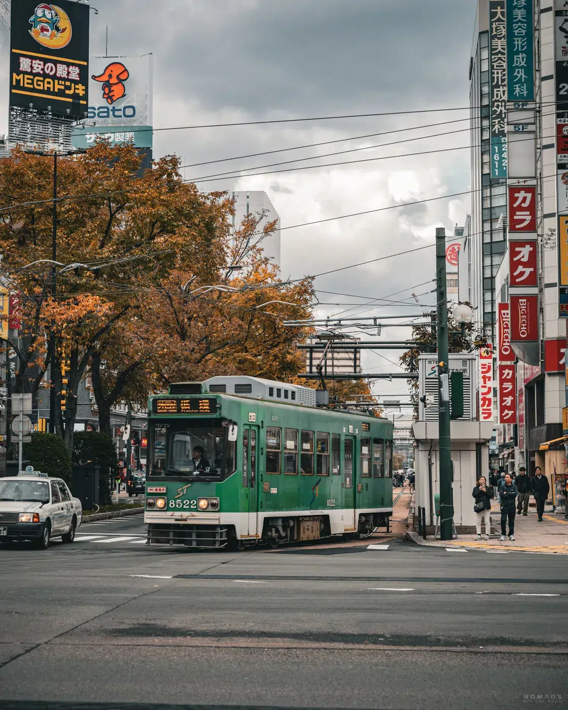 Grüne Straßenbahn in der Innenstadt von Sapporo im Herbst, umgeben von buntem Laub und japanischen Reklametafeln.