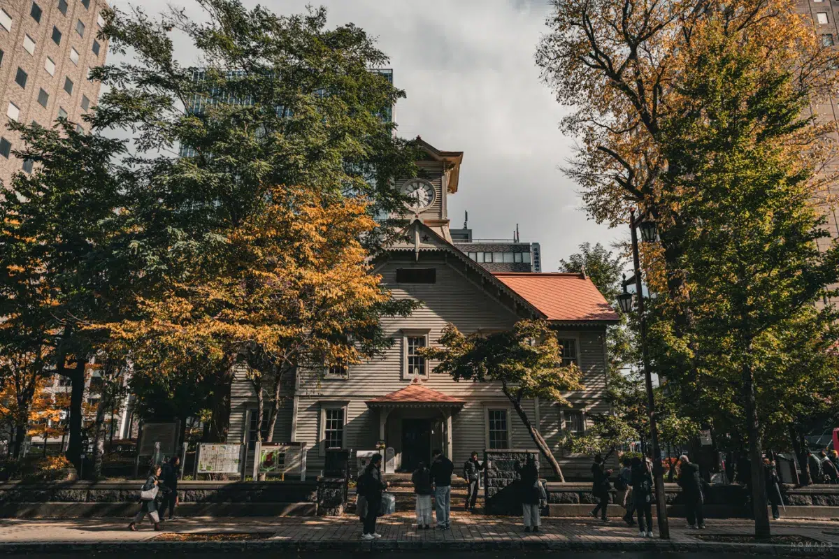 Außenansicht des Sapporo Clock Tower inmitten herbstlicher Bäume, mit Besuchern vor dem Eingang.