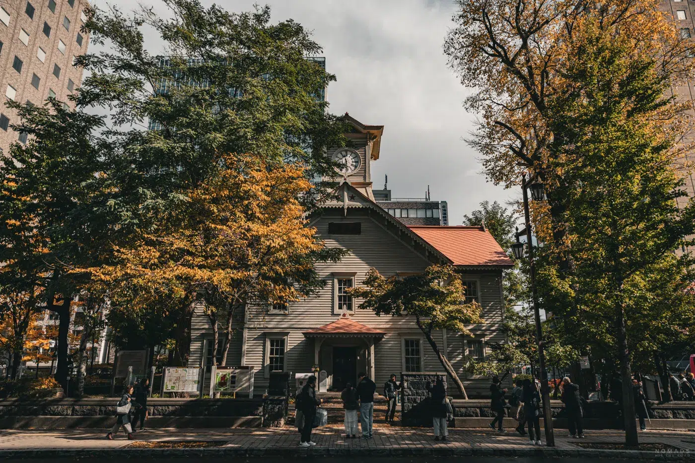 Außenansicht des Sapporo Clock Tower inmitten herbstlicher Bäume, mit Besuchern vor dem Eingang.