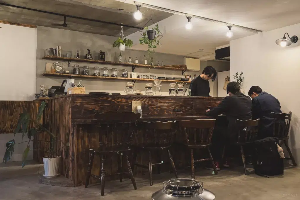 Gemütliches, minimalistisches Café mit Holztheke und Hockern, in dem ein Barista Kaffee zubereitet, während zwei Männer in Anzügen an der Theke sitzen.