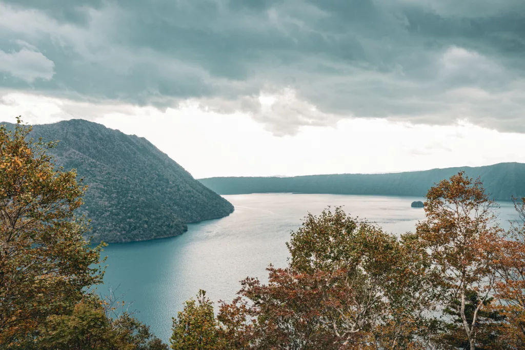 Blick auf den tiefblauen Lake Mashu auf Hokkaido, Japan – umgeben von bewaldeten Berghängen und Herbstlaub unter dramatisch bewölktem Himmel.