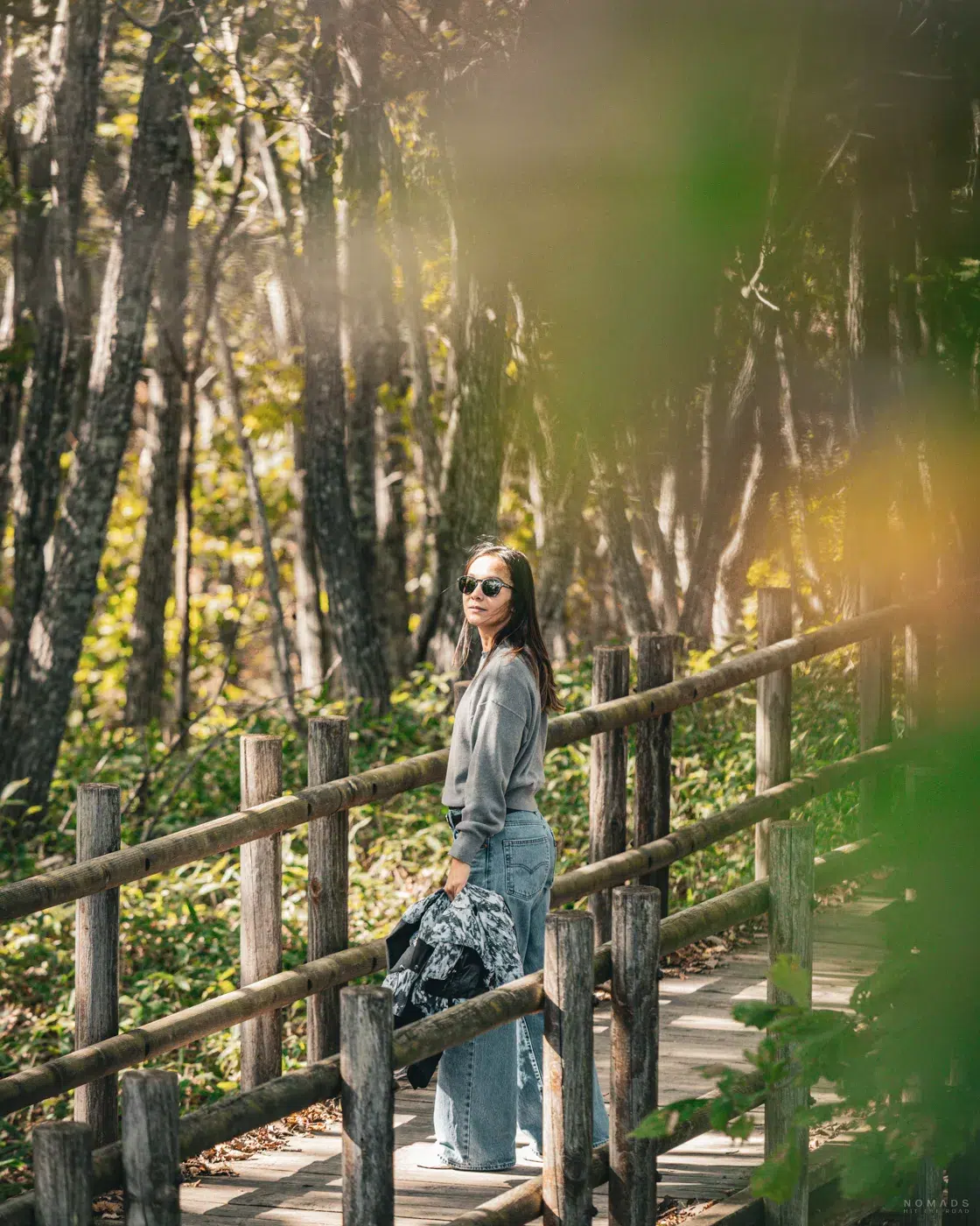Frau auf einem schmalen Holzsteg im lichten Wald bei Kushiro, umgeben von Sonnenlicht und herbstlichem Blattgrün.