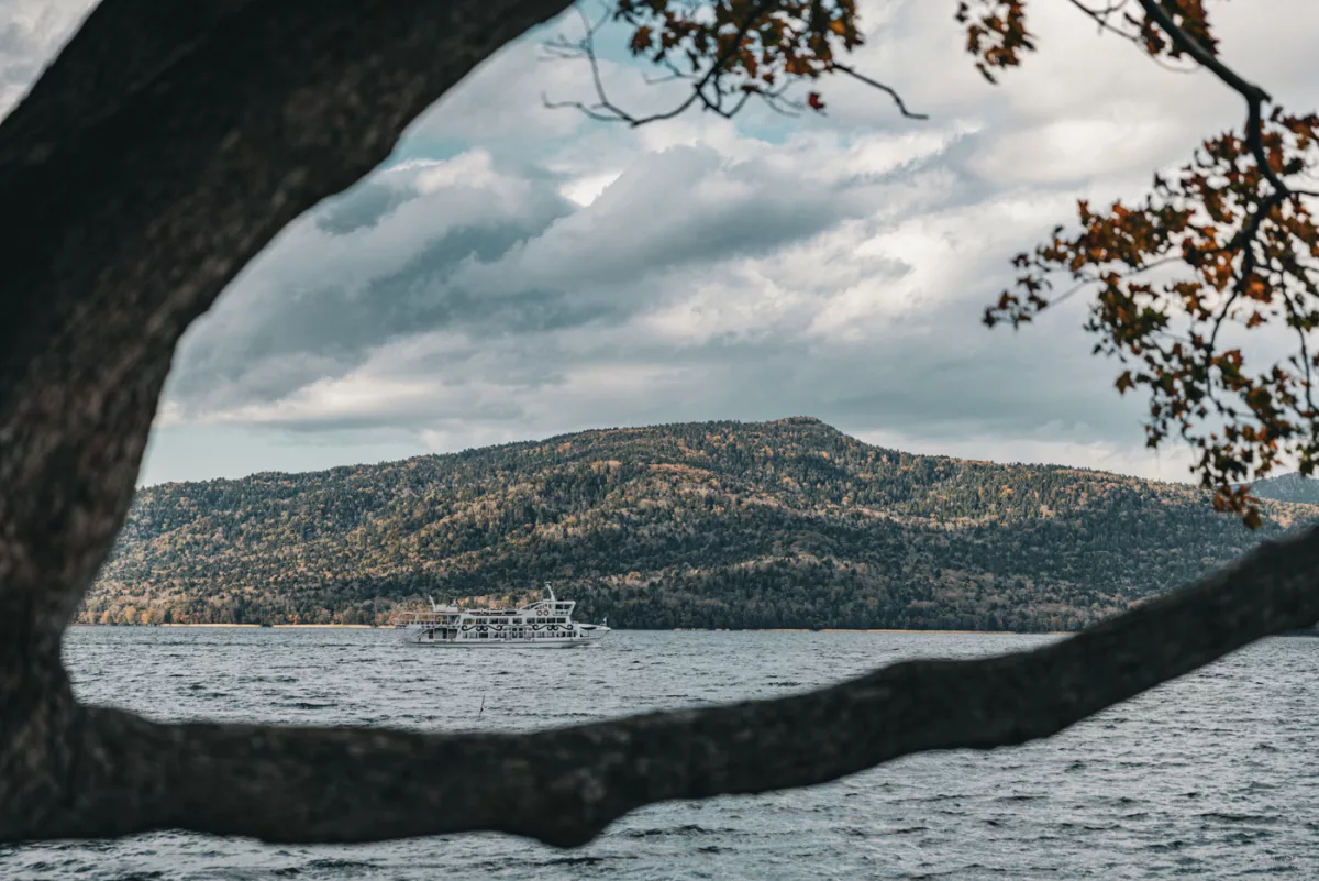 Fähre auf dem Lake Akan, eingerahmt von Ästen, mit bewaldeten Bergen im Hintergrund.
