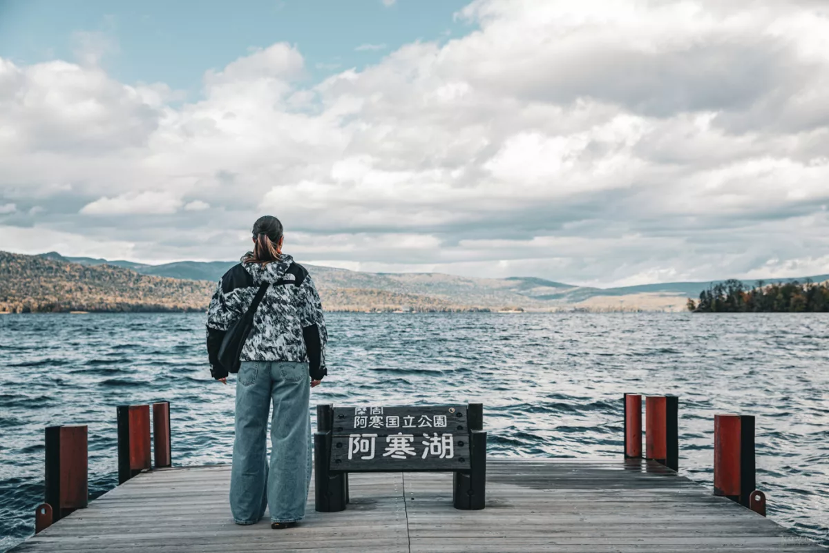 Person steht auf einem Holzsteg am Lake Akan und blickt über den windigen See in die Vulkanlandschaft.