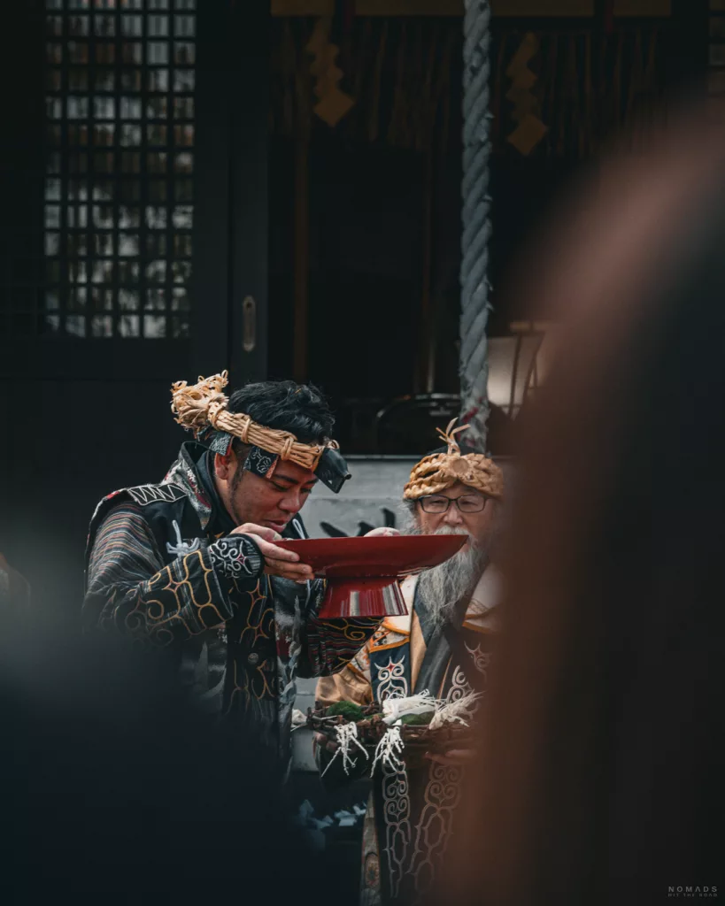 Mann trinkt aus einer roten zeremoniellen Schale, während ein älterer Ainu-Mann daneben eine Opfergabe hält – beide tragen verzierte Roben und Kopfbedeckungen.