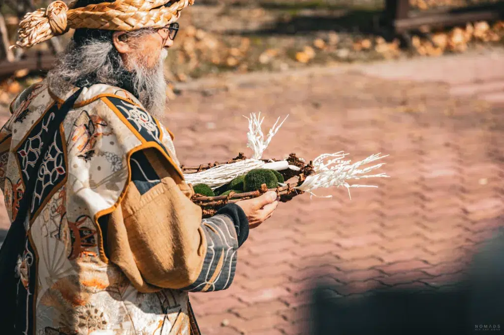 Ainu-Ältester mit traditioneller Kleidung hält eine mit weißen Ritualstäben und Marimo-Algen geschmückte Opfergabe.