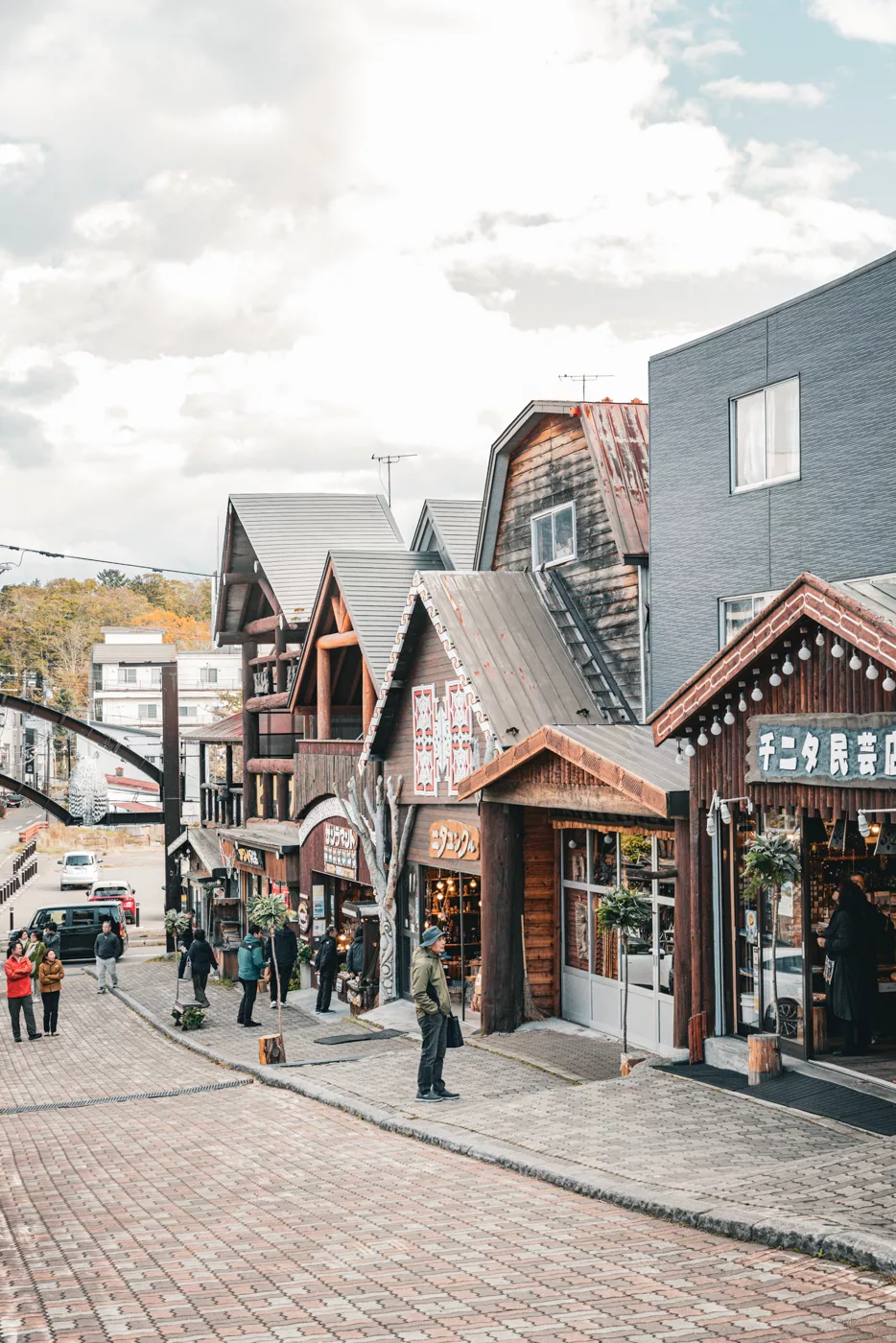 Kopfsteinpflasterstraße in Akan mit traditionellen Holzhäusern im Ainu-Stil, gesäumt von Touristen und dekorativen Geschäften.