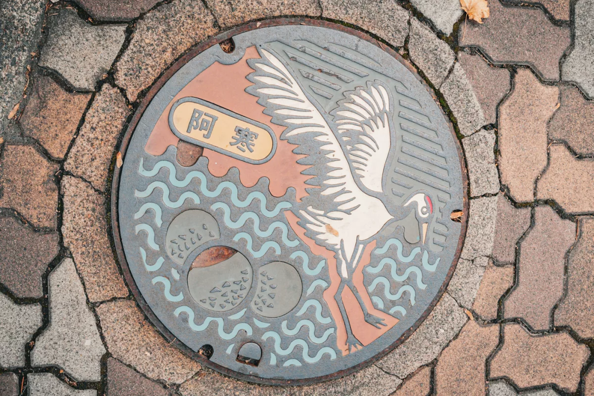Bunter Gullydeckel in Akan, Hokkaido, mit Kranich, Wellen, Bergen und japanischer Schrift – Symbolik der Region in kunstvollem Design.