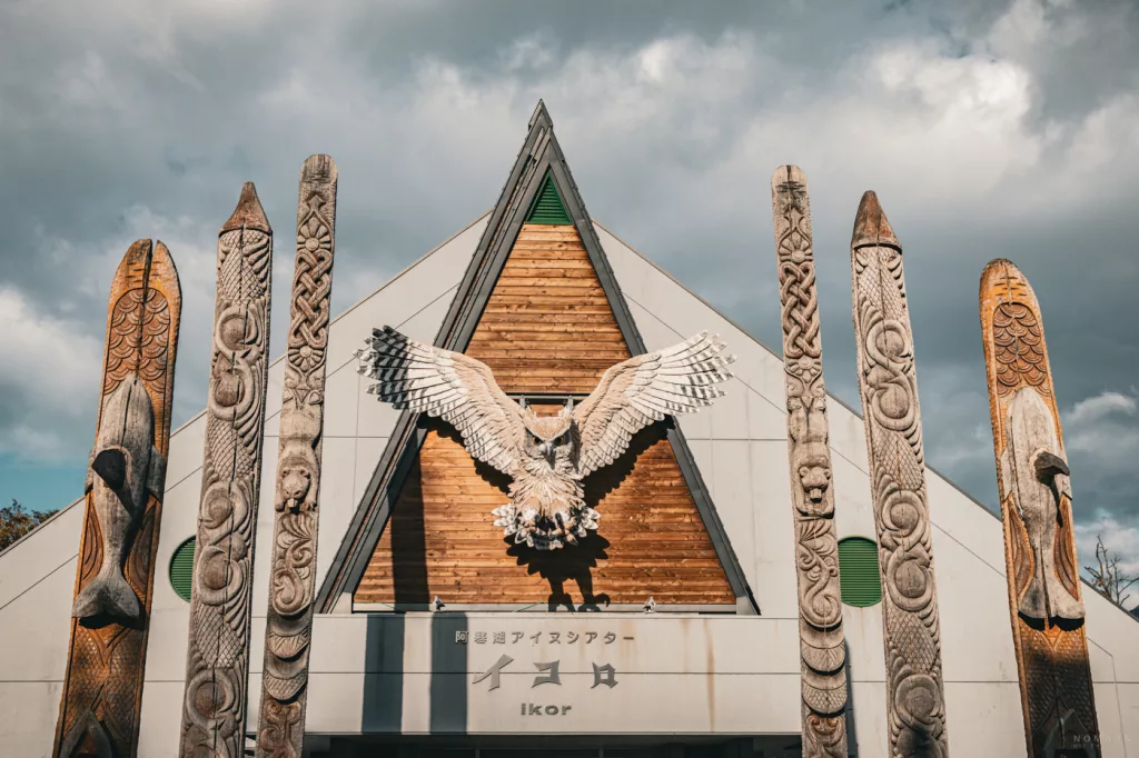 Fassade des Ainu-Theaters Ikor mit imposanter Eulen-Skulptur über dem Eingang, umrahmt von kunstvoll geschnitzten Holzpfählen mit Tier- und Naturmotiven.