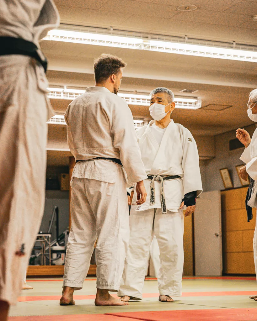 Ein älterer japanischer Judomeister im weißen Gi und mit schwarzem Gürtel spricht mit einem jüngeren westlichen Judoka während einer Trainingseinheit in einer traditionellen Dojo-Halle.