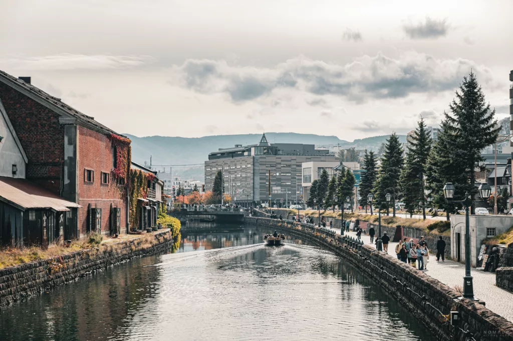 Blick über den Otaru Canal mit herbstlichen Farben, alten Lagerhäusern und einem Ausflugsboot auf dem Wasser.