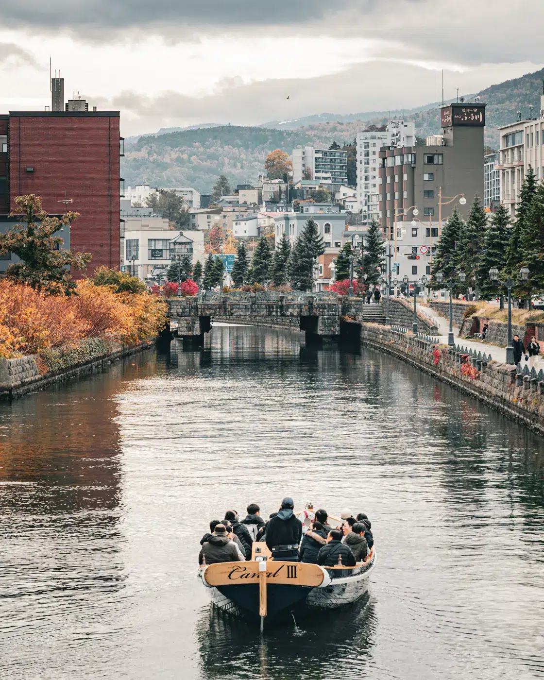 Bootstour auf dem Otaru-Kanal im Herbst, umgeben von historischen Lagerhäusern und buntem Laub, mit Blick auf die Stadt und die umliegenden Berge.