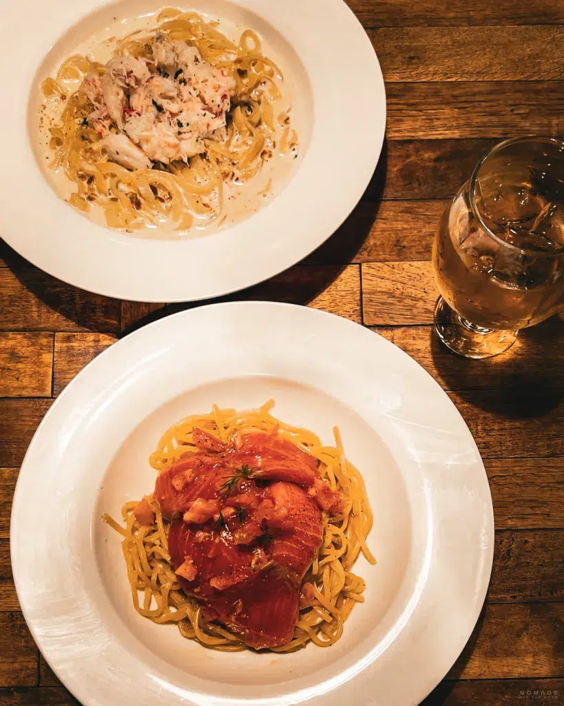 Zwei Pastagerichte in weißen Tellern auf einem Holztisch serviert – oben eine cremige Pasta mit Krabbenfleisch, unten ein Teller mit Lachs-Pasta, daneben ein Glas Eistee.