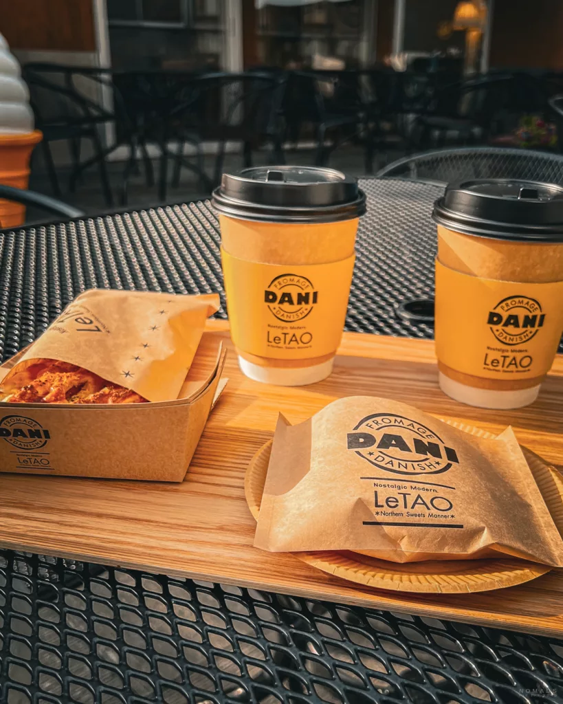 Tablett mit Kaffee in gelben LeTAO-Bechern und dänischem Gebäck in Papierverpackung auf einem schwarzen Metalltisch im Außenbereich eines Cafés.