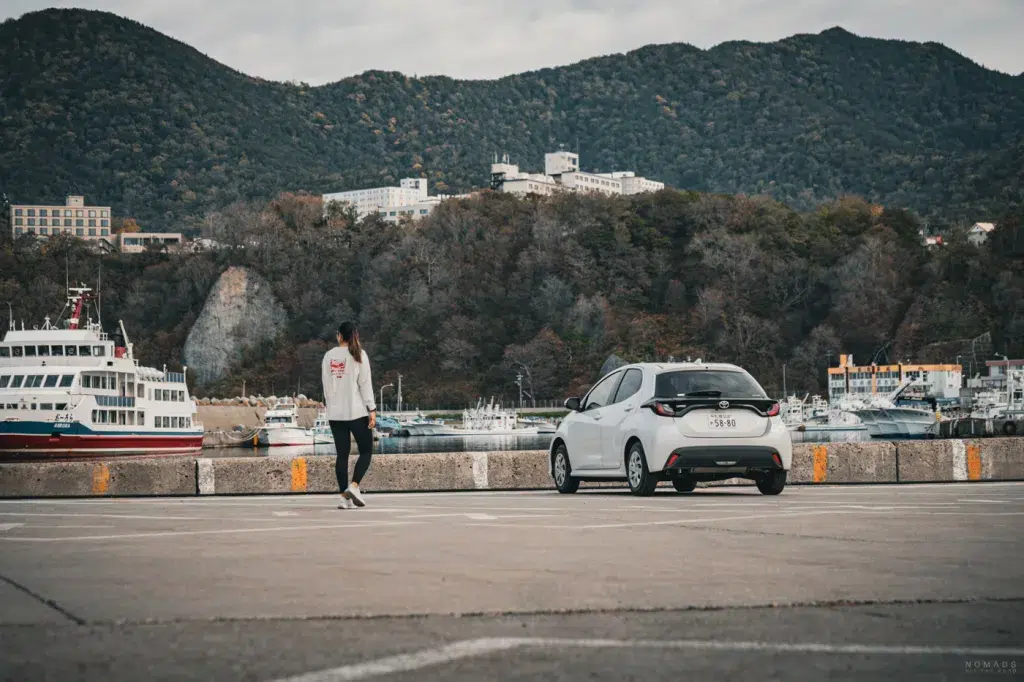 Reisende mit Mietwagen am Hafen auf Hokkaido – Blick auf Boote, Küste und bewaldete Hügel.