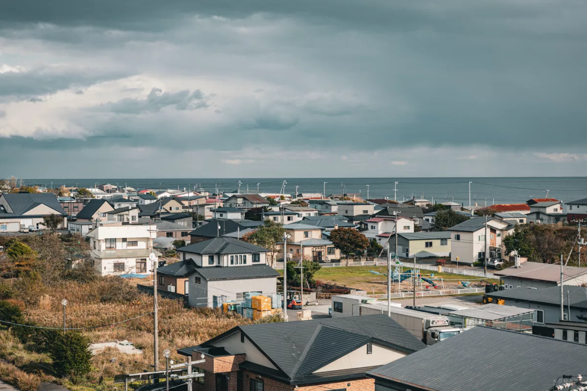 Blick über den Küstenort bei Noboribetsu mit Wohnhäusern, Spielplatz und dem Meer im Hintergrund unter dramatischem Himmel.