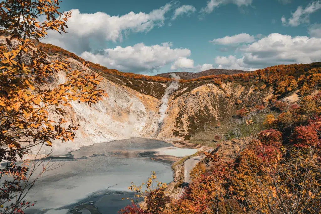 Blick auf den türkisfarbenen Oyunuma-See bei Noboribetsu, umgeben von herbstlich gefärbten Hügeln und dampfenden Fumarolen unter blauem Himmel.