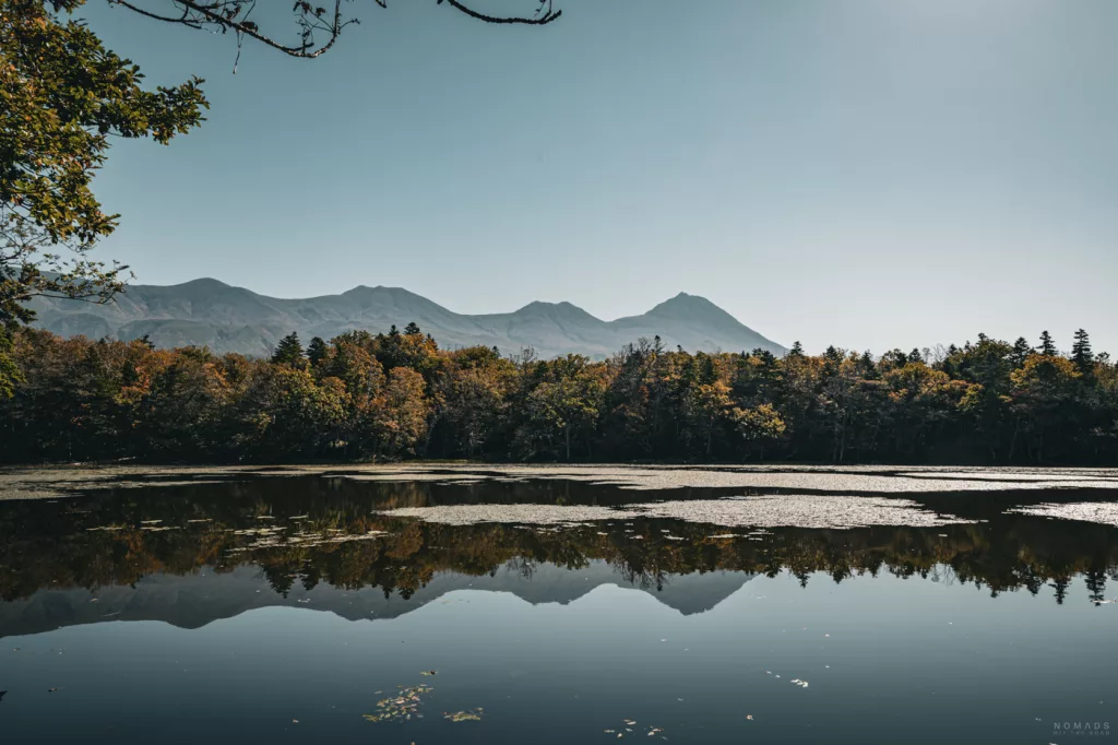 Blick über einen der Shiretoko Five Lakes mit ruhiger Wasseroberfläche, die herbstlich gefärbte Wälder und die Bergkette des Shiretoko-Gebirges unter klarem Himmel spiegelt.