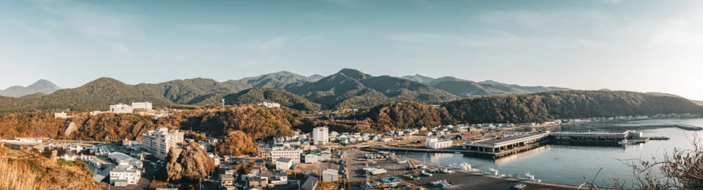 Panoramablick über Utoro und den Hafen von Shiretoko mit Booten, Gebäuden am Wasser und bewaldeten Hügeln im Hintergrund bei warmem Licht.