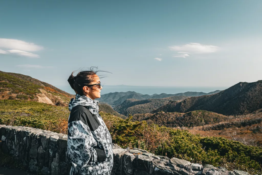 Frau steht am Aussichtspunkt am Shiretoko Pass und blickt über die bewaldeten Berge bis zum Meer im Hintergrund, herbstliche Landschaft auf Hokkaido.