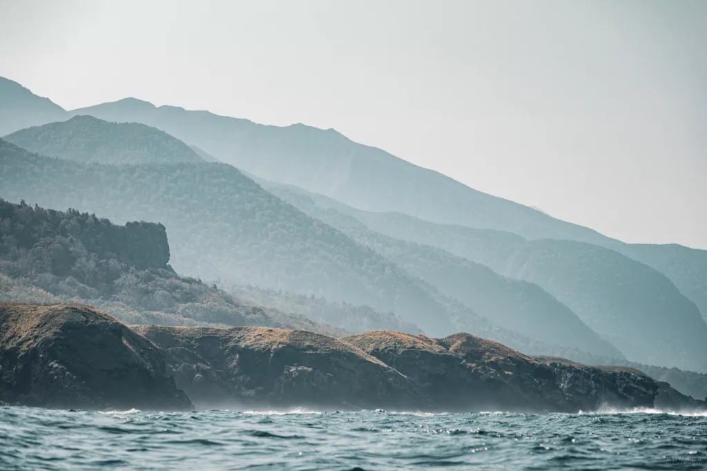Küstenlandschaft am Kap Shiretoko: Blick vom Boot auf zerklüftete Felsen, bewaldete Berghänge und das offene Meer unter leichtem Dunst.