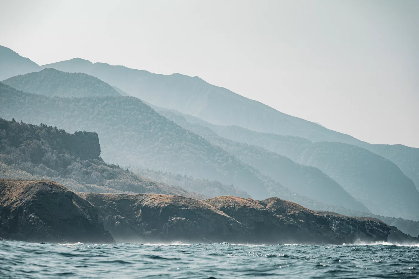 Küstenlandschaft am Kap Shiretoko: Blick vom Boot auf zerklüftete Felsen, bewaldete Berghänge und das offene Meer unter leichtem Dunst.