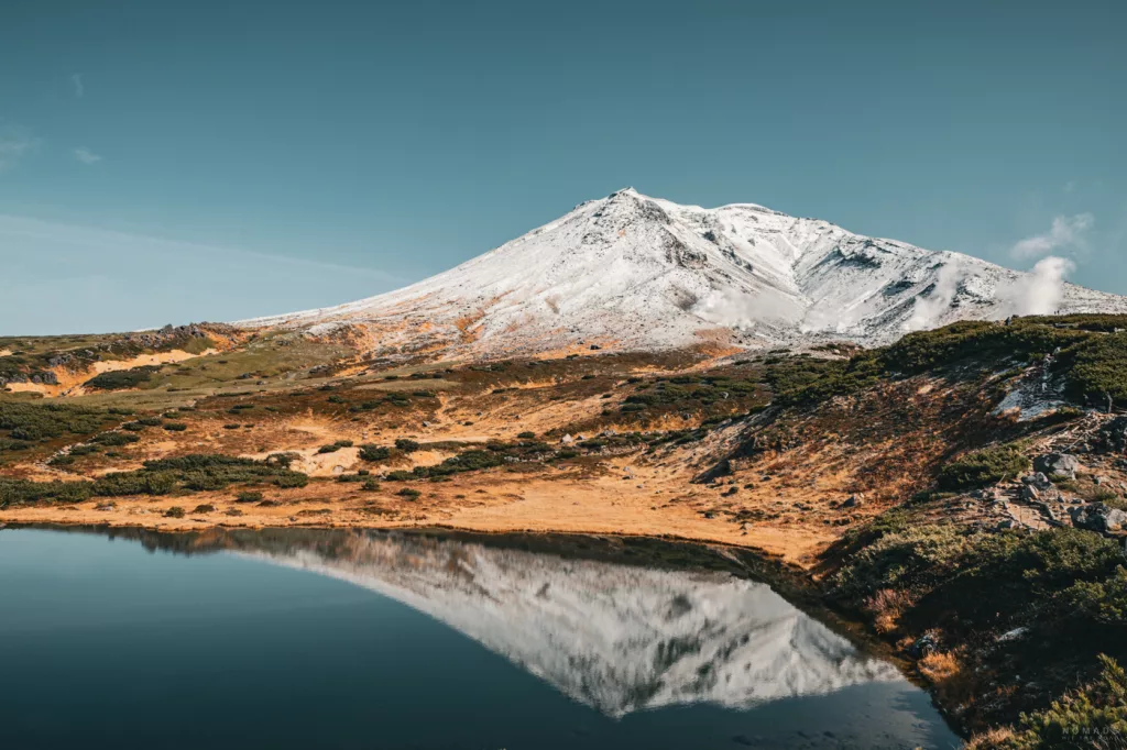Asahidake im Daisetsuzan National Park auf Hokkaido mit schneebedecktem Gipfel, Schwefeldampf und Spiegelung im Sugatami-See im Herbst