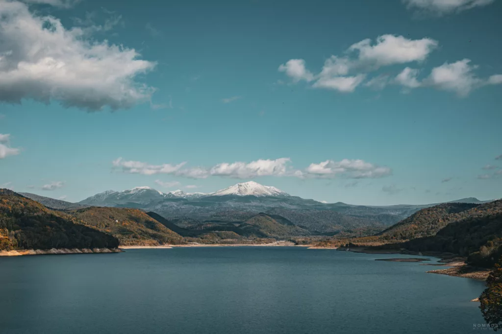 Weitblick über einen ruhigen See in Hokkaido mit schneebedecktem Asahidake im Hintergrund, umgeben von herbstlich gefärbten Hügeln und blauem Himmel mit vereinzelten Wolken.