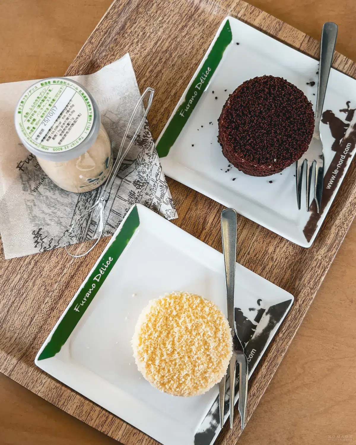 Zwei kleine Küchlein – ein dunkles Schokoladendessert und ein helles, mit Bröseln bestreutes Küchlein – auf weißen Tellern mit der Aufschrift „Furano Delice“, daneben ein Glas Dessert auf einem Holztisch.