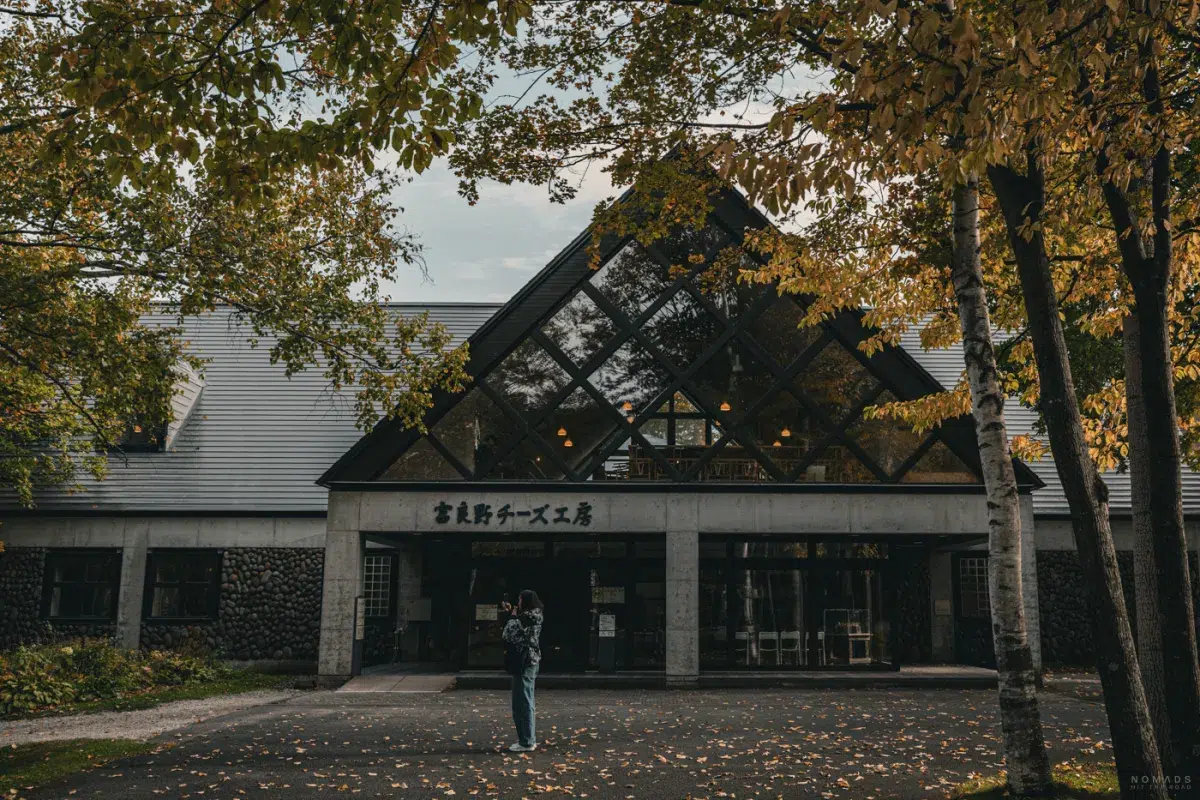 Eingang der Furano Cheese Factory mit markanter Glasfassade, umgeben von herbstlichen Bäumen in Furano, Hokkaido