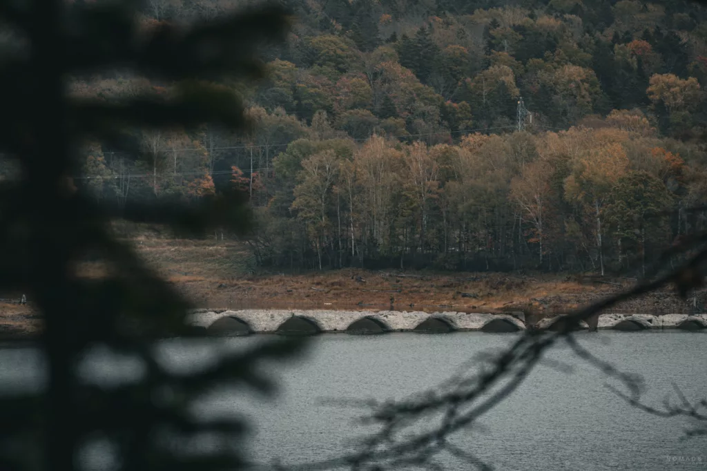 Steinbogenbrücke (Taushubetsu Bridge) am Ufer eines Sees in Hokkaido, umgeben von herbstlich gefärbtem Wald und leicht unscharfen Ästen im Vordergrund.