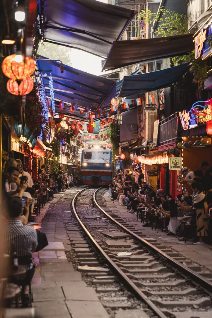Zug fährt auf einer Gleise auf der Hanoi Train Street während rechts und links Menschen in Cafés sitzen